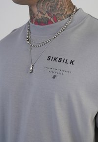 Halljahuuline hall t-särk lühikeste varrukatega, must "SIKSILK" logo, kihilised hõbedased kaelakeed ja tekstuuriga võlu ripatsiga detail.