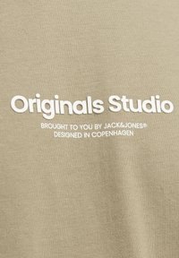 Khakifarbendes Baumwoll-T-Shirt mit weißem Drucktext: "Originals Studio," "präsentiert von JACK&JONES," und "entworfen in Kopenhagen."