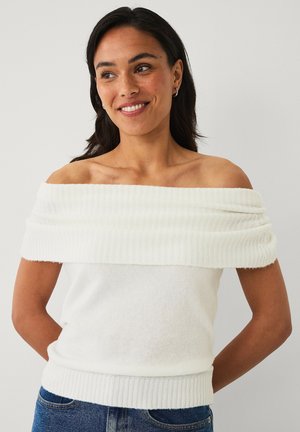 Vrouw met donker haar lacht en kijkt opzij, draagt een witte gebreide off-shoulder top en blauwe spijkerbroek tegen een effen achtergrond.