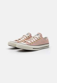 Pink canvas sneakers with white laces, gold eyelets, and black rubber sole. Features a textured upper and striped detailing on the sole.