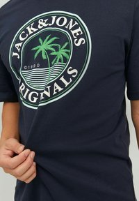 Mörkblå bomulls-t-shirt med en cirkulär logotyp med palmer, texten 'JACK & JONES ORIGINALS' och en randig bakgrundsdesign.