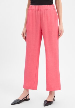 Femme portant un pantalon ample couleur corail rose avec une taille élastique et des chaussures slingback noires à bout pointu sur fond blanc.