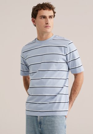 Muž v svetlomodrom krátkom tričku s tenkými čiernymi horizontálnymi pruhmi a svetlomodrých džínsoch, stojaci s rukami za chrbtom.