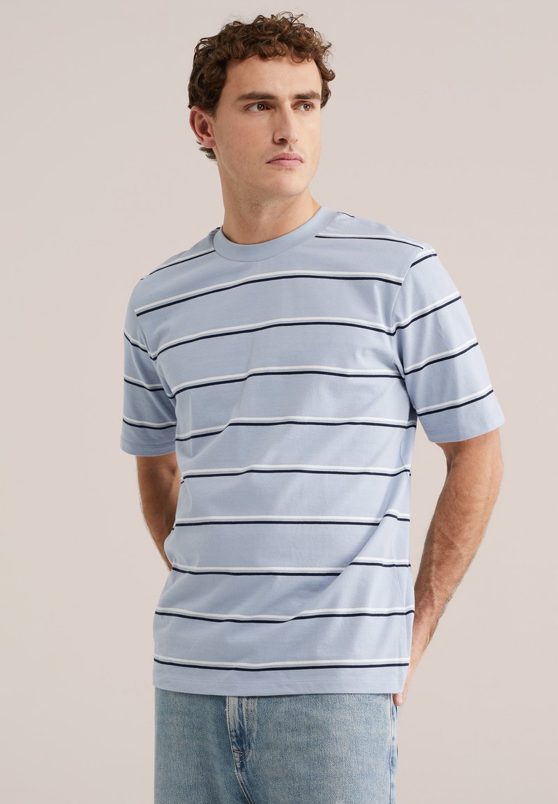 Bărbat purtând tricou cu mâneci scurte, albastru deschis, cu dungi subțiri orizontale negre și blugi albastru deschis, stând cu mâinile la spate.