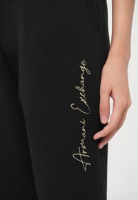 Mustat jogginghousut pehmeästä materiaalista, joissa on kultaisella langalla kirjotettu "Armani Exchange" -teksti sivussa ja muotoiltu malli.