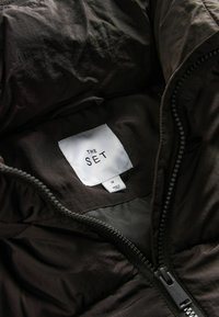 Intérieur de veste isolante noire avec une texture lisse, arborant une étiquette blanche avec l'inscription "THE SET", taille M, et une fermeture éclair.