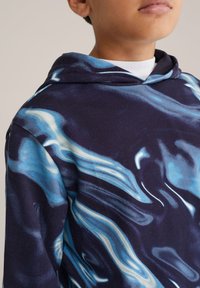 Kapucna majica v temno mornarsko modri barvi z vrtinčastimi modrimi in belimi abstraktnimi vzorci. Tkanina je videti mehka z gladko teksturo.