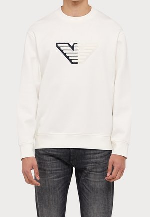 Homme portant un sweat-shirt blanc à manches longues avec un grand logo ailé noir et blanc sur la poitrine et un jean foncé.