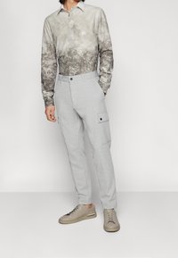 Koszula na guziki w marmurkowy wzór w odcieniach szarości i beżu, zestawiona z jasnoszarymi spodniami typu cargo z bocznymi kieszeniami i czarnymi akcentami. Jasne obuwie.