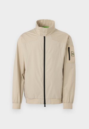 Beige leichte Jacke mit Stehkragen, schwarzem Frontreißverschluss, elastischen Bündchen und Reißverschlusstasche am linken Ärmel mit Logoaufnäher.