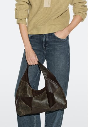 Person trägt einen beigen Pullover und blaue Jeans und hält mit einer Hand eine dunkelbraune Patchwork-Ledertasche am Griff.