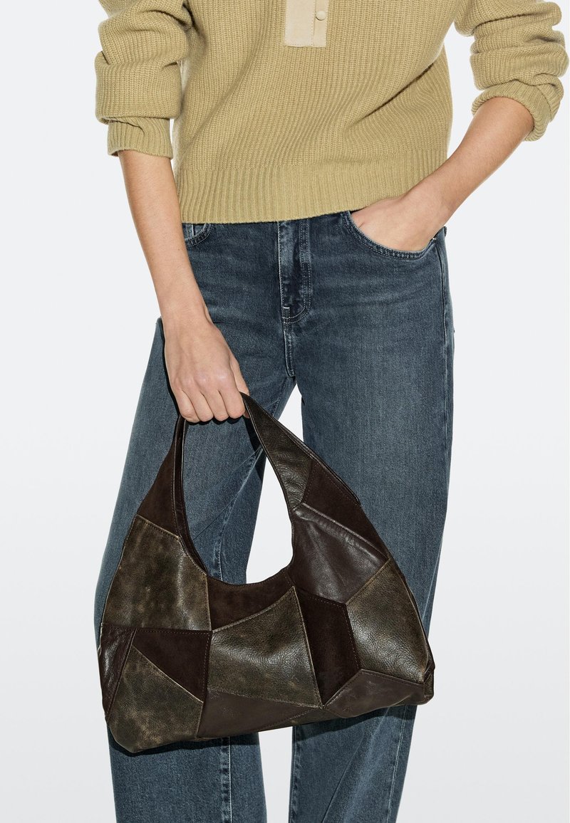 Person trägt einen beigen Pullover und blaue Jeans und hält mit einer Hand eine dunkelbraune Patchwork-Ledertasche am Griff.