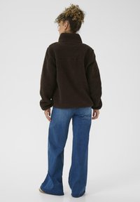 Pullover in pile marrone con colletto alto, caratterizzato da una superficie morbida e texturizzata. Abbinato a jeans a gamba larga di colore blu. Vestibilità e design standard.