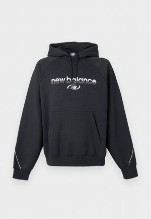 Νέος άνδρας με μαύρο φούτερ New Balance με λογότυπο στο στήθος και σκουρόχρωμο γκρι παντελόνι, στέκεται μπροστά σε απλό φωτεινό φόντο.