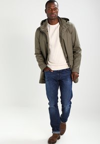 Chaqueta de lluvia color caqui con capucha, sobre un suéter de punto de cable crema, combinada con jeans azules y botas de tobillo marrones. Diseño simple y funcional.