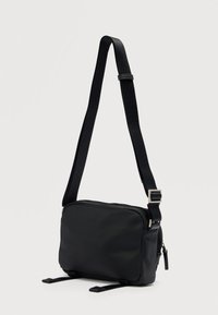 Μαύρη τσάντα ώμου από μαλακό ύφασμα. Διαθέτει μακριά ρυθμιζόμενη τιράντα και κλείσιμο με φερμουάρ, με πλαϊνές λεπτομέρειες και μινιμαλιστικό σχεδιασμό.