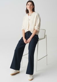 Creme gebreide cardigan met knopen, gecombineerd met donkerblauwe wijde broek en witte sneakers. Soepele stof en ontspannen pasvorm.