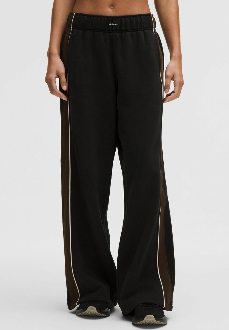 Zwarte wijde sweatpants met bruine zijpanelen en witte bies, gedragen met zwarte sneakers, elastische tailleband met klein wit logo.