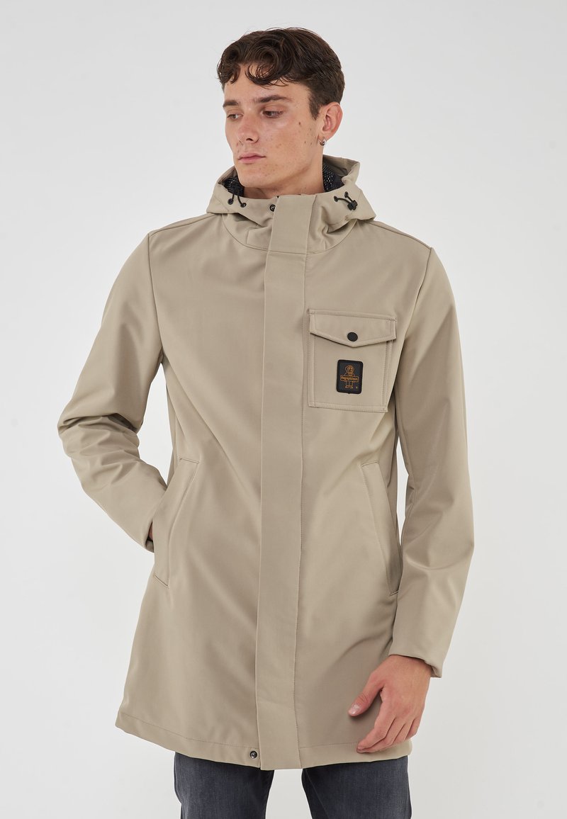 Cappotto impermeabile beige con cappuccio, tasca sul petto e tasche laterali. Tessuto liscio con chiusura a scatto frontale e dettaglio con cordino.