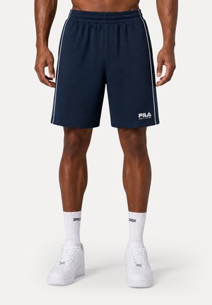 Homme portant un short FILA bleu marine avec des rayures blanches, des chaussettes blanches et des baskets blanches, debout devant un fond uni.