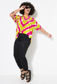Helles Batik-Shirt mit grünen und rosa Mustern, schwarze Cargo-Hosen und schwarze Sandalen mit Riemen. Model trägt Sonnenbrille und posiert.