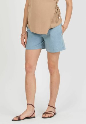Kvinde iført lyseblå jeansshorts, beige top med omslagsstil, brune flade sandaler med stropper og gyldne armbånd på det ene håndled.