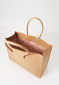 Sac fourre-tout en cuir tan avec un extérieur texturé et un intérieur lisse. Il dispose de deux poignées et d'une poche intérieure, avec un logo embossé à l'avant.