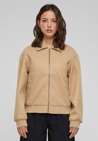 Beige zip-jakke med flat krage, ribbet kant og teksturert stoff. Har sidelommer og avslappede ermer.