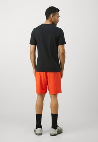 T-shirt noir à manches courtes associé à un short orange, complété par des chaussettes noires et des chaussures de sport grises ; le tissu semble lisse et ajusté.