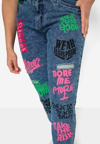 Vaqueros de mezclilla con múltiples estampados gráficos en rosa, verde y negro, que presentan frases como "Luce con orgullo los vaqueros" y "Asume el riesgo". Presenta zonas desgastadas.