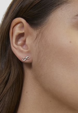 Silberne Steckohrringe in Form eines Unendlichkeitssymbols, mit kleinen klaren Steinen entlang einer Kante, vor hellem Hautton.