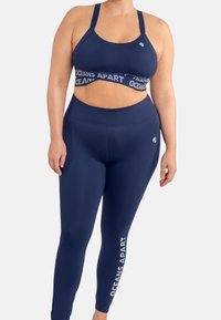 Sportivo blu con ampia fascia elastica etichettata "OCEANS APART", abbinato a leggings a vita alta che presentano lo stesso testo sulla gamba.
