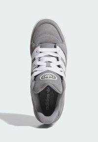 Gray sneaker with suede and leather materials, featuring white laces, three white stripes, and a round logo on the tongue. Flat sole design.