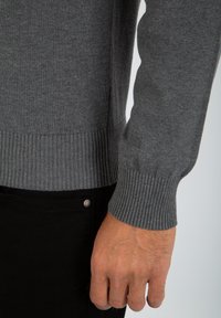 Maglione grigio lavorato a maglia con polsini e orlo a coste, realizzato in materiale morbido. Il braccio visibile appare ben aderente, enfatizzando texture e dettagli di design.