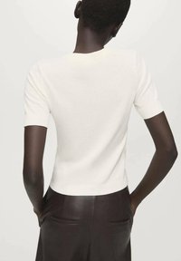 Bela pletena majica s kratkimi rokavi ima oprijet kroj z okrolim izrezom in rebrastim robom na rokavih ter spodnjem delu. Kombinirana s temnimi hlačami.