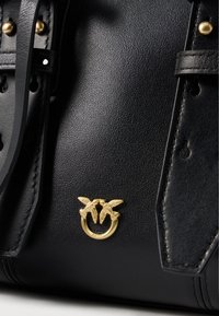 Melnas ādas rokassomiņa ar dekoratīvu zelta aparatūru, kas veidota divu putnu un apļa akcenta formā, ar teksturētu virsmu un siksnu detaļām.