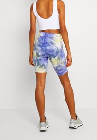 High-waisted, tie-dye workout shorts in blue and yellow, made of stretchy fabric, paired with a white fitted tank top and gray sneakers.