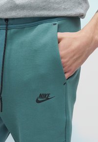 Χέρι στην τσέπη πράσινου παντελονιού φόρμας Nike με μαύρο κορδόνι, φορεμένο με γκρι μπλουζάκι ορατό στη μέση.