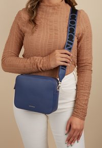 Modrá texturovaná kožená crossbody taška s odnímatelným modrým páskem s černým nápisem. Čtvercový tvar se stříbrnými kovovými doplňky.