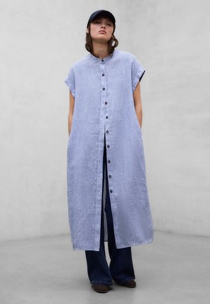 Kobieta ubrana w długą, jasnoniebieską, bezrękawnikową sukienkę zapinaną na guziki, na ciemnych spodniach, w czapce z daszkiem i brązowych butach, stojąca na tle szarej ściany.