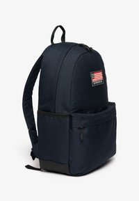 Mochila de tela azul marino con forma redondeada, que cuenta con un bolsillo frontal, parche con el logo y bandera americana, y acentos en negro en la parte inferior.