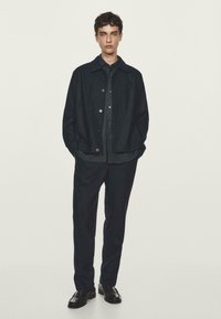 Giacca di jeans scura con chiusura a bottoni, indossata sopra una camicia a righe scure e abbinata a pantaloni scuri su misura. Un paio di scarpe in pelle nere completa il look.