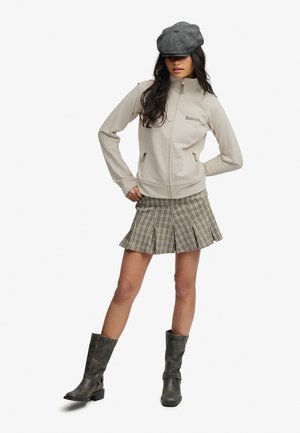 Femme portant une veste beige zippée, une jupe plissée à carreaux, une casquette gavroche grise et des bottes noires mi-mollet, debout les mains sur les hanches.