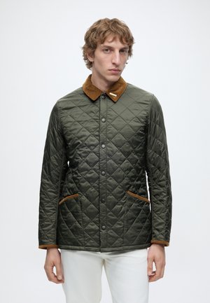 Homme portant une veste matelassée verte avec un col en velours côtelé marron et un bord de poche, debout devant un fond blanc uni.