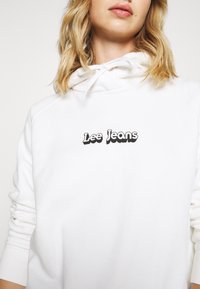 Sudadera blanca con capucha, con un logo negro de "Lee Jeans" en un tipo de letra en negrita. La tela tiene una textura suave y puños de canalé.
