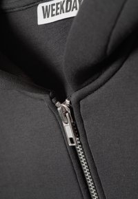 Black zip-up hoodie featuring a silver zipper, soft fabric, and a label with "WEEKDAY" text inside the collar area.