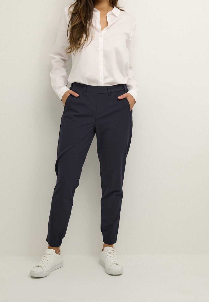 Femme portant une chemise blanche boutonnée, un pantalon bleu marine fuselé avec les mains dans les poches, et des baskets blanches, debout devant un fond clair uni.