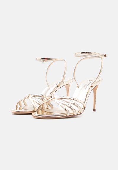 Ζευγάρι μεταλλικές χρυσές σανδάλες με ψηλό τακούνι, λουριά αστραγάλου και πολλά λεπτά μπροστινά λουριά, τοποθετημένες σε απλό φωτεινό φόντο.