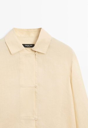 Beżowa koszula z długim rękawem, z ostrym kołnierzem i ukrytym listwą guzikową z trzema widocznymi guzikami.