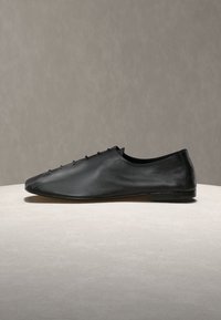 Μαύρο δερμάτινο παπούτσι με κομψό, μύτη σχεδιασμό. Λεπτό υφή, ελάχιστα κορδόνια στο πάνω μέρος και επίπεδη σόλα χωρίς ορατά μεταλλικά στοιχεία.